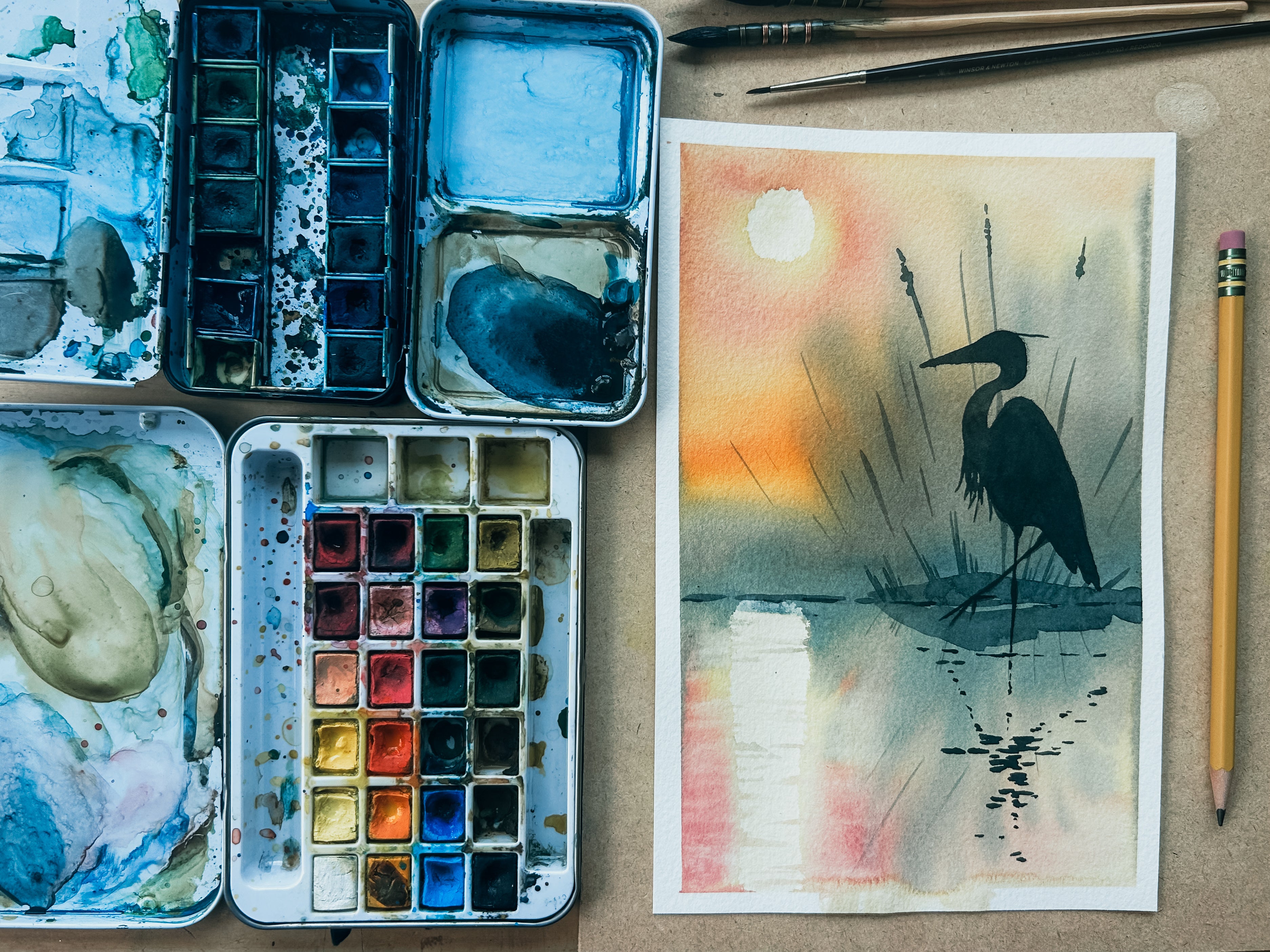 Watercolor painting of a bird on a piece of paper with watercolor pans and brushes on a table.