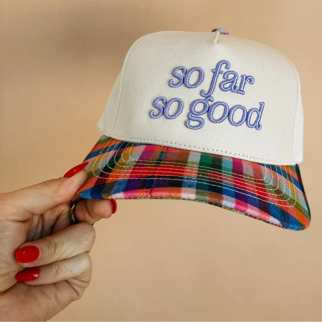 A hand with red nail polish holds a white snapback cap featuring blue embroidered text that reads 'so far so good' on the front. The brim has a vibrant, multicolored plaid pattern.