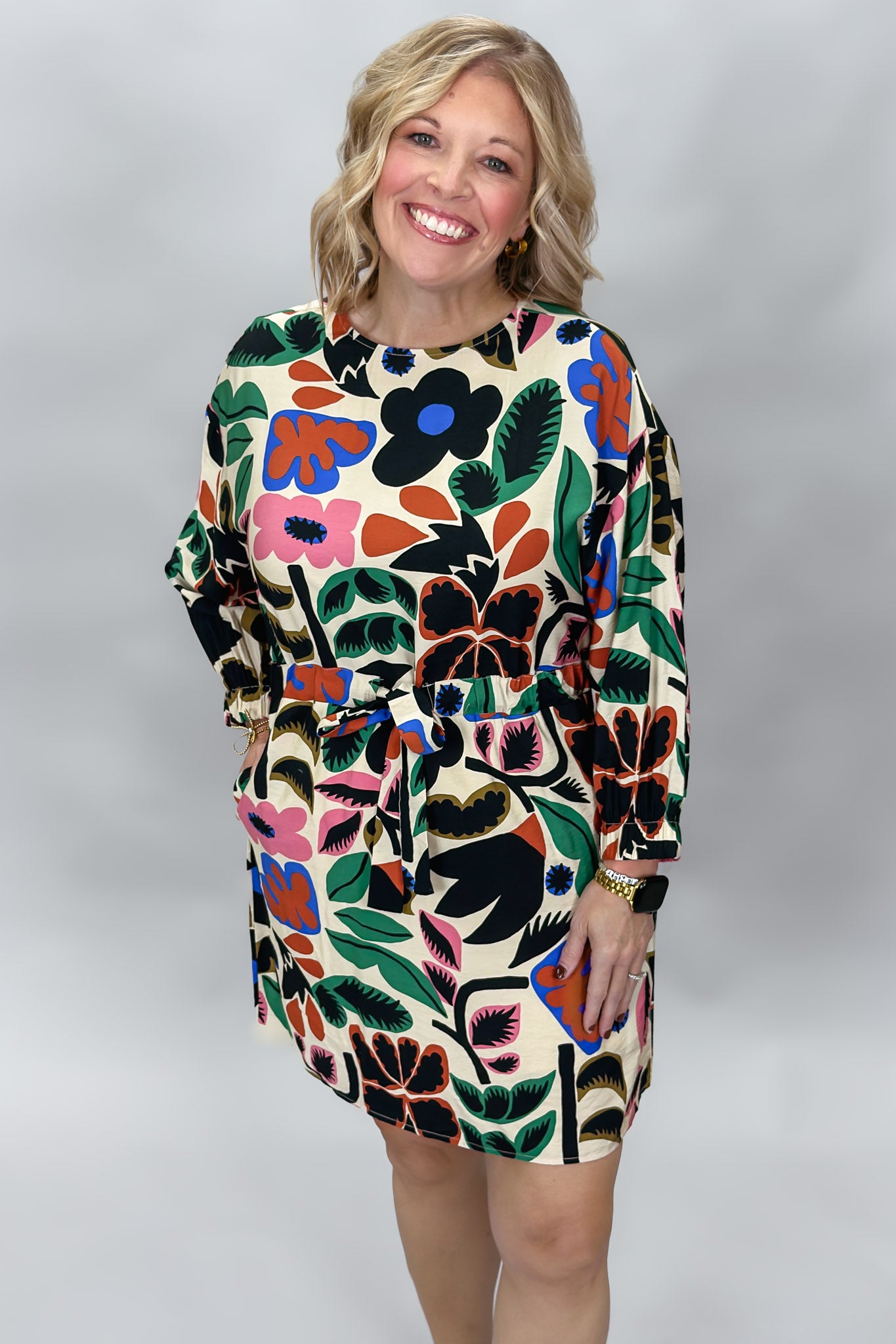 Three-quarter front view of the Back On 74 bold floral print midi dress on a model, highlighting its belted silhouette, long sleeves, knee-length cut, and large colorful floral print against cream.