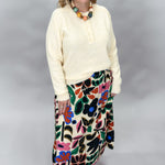 Candle Flame bold floral print midi skirt with large multicolor flowers and leaves on cream, styled with a cream knit pullover, beaded necklace, and tan sandals; full front view on a model.