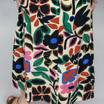 Rear waist-to-hem view of the Candle Flame bold floral print skirt, featuring the cream-ground multicolor botanical pattern, knee-length A-line silhouette, and brown wedge sandals.