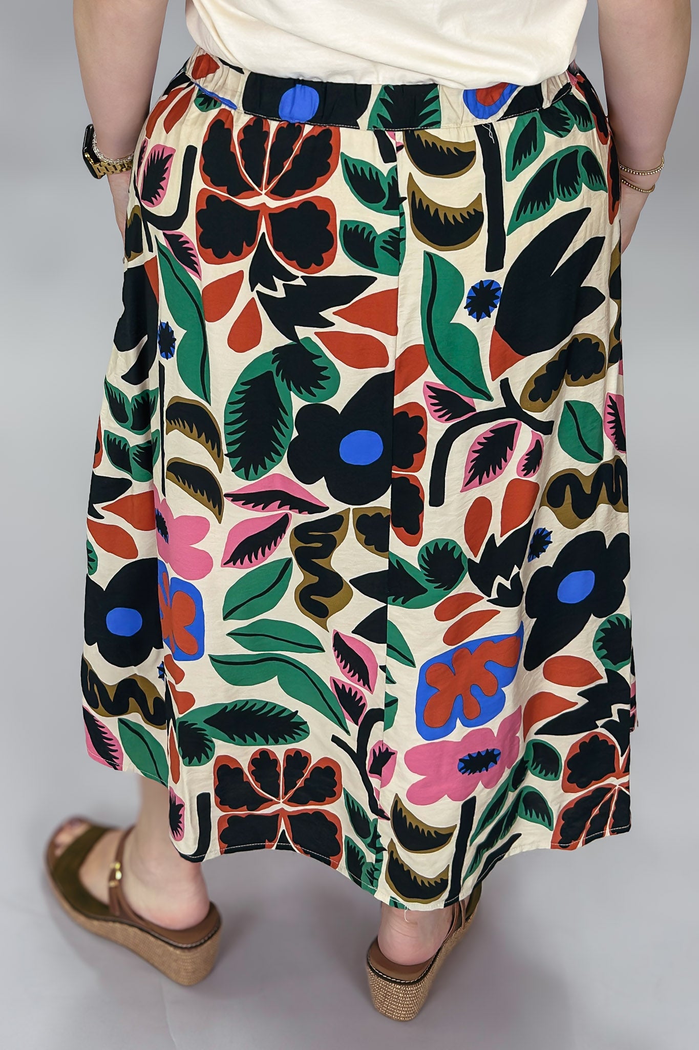 Rear waist-to-hem view of the Candle Flame bold floral print skirt, featuring the cream-ground multicolor botanical pattern, knee-length A-line silhouette, and brown wedge sandals.