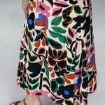 Candle Flame midi skirt close-up from waist to hem, showing the high-rise A-line cut, bold oversized multicolor floral and leaf print on cream, and the model’s brown wedge sandals.