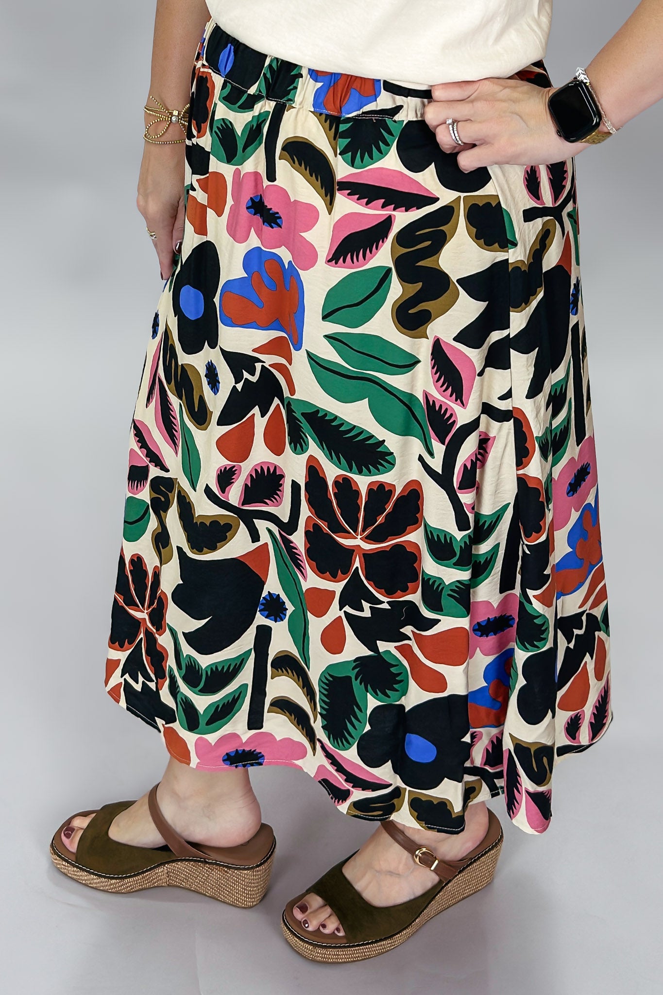 Candle Flame midi skirt close-up from waist to hem, showing the high-rise A-line cut, bold oversized multicolor floral and leaf print on cream, and the model’s brown wedge sandals.