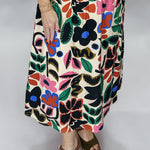 Close view of the Candle Flame bold floral skirt, showing its cream-ground multicolor flower and leaf pattern, knee-length A-line shape, and drape with brown wedge sandals.