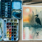 Watercolor painting of a bird on a piece of paper with watercolor pans and brushes on a table.
