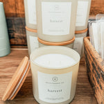 A jar candle labeled 'harvest' by Hico Candle Co. sits on a wooden shelf with its wooden lid beside it.