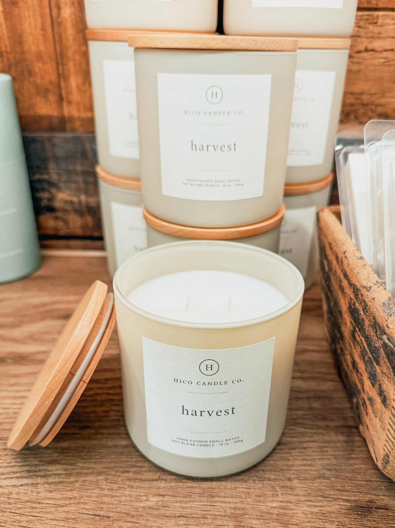 A jar candle labeled 'harvest' by Hico Candle Co. sits on a wooden shelf with its wooden lid beside it.
