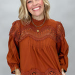 A smiling blonde woman wearing a rust-orange long-sleeve blouse with intricate lace trim at the neckline and hem, paired with brown and black plaid pants, against a solid gray studio background.