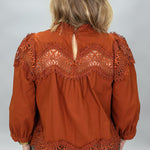 A woman viewed from behind wearing a rust-orange blouse with intricate lace detailing across the shoulders and hem, paired with plaid pants, against a light gray studio background.