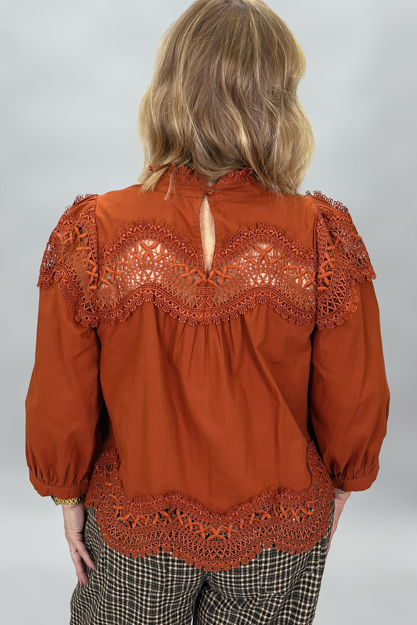 A woman viewed from behind wearing a rust-orange blouse with intricate lace detailing across the shoulders and hem, paired with plaid pants, against a light gray studio background.