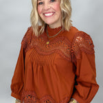 A smiling blonde woman wearing a rust-orange long-sleeve blouse with intricate lace trim at the neckline, shoulders, and hem, paired with plaid pants, against a neutral gray studio background.