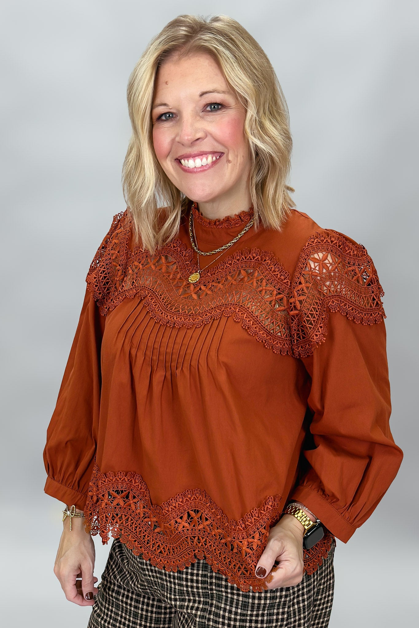 A smiling blonde woman wearing a rust-orange long-sleeve blouse with intricate lace trim at the neckline, shoulders, and hem, paired with plaid pants, against a neutral gray studio background.