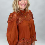 A smiling blonde woman wearing a rust-orange long-sleeve blouse with intricate lace trim at the neckline, cuffs, and hem, paired with brown and black plaid pants, against a neutral gray studio background.