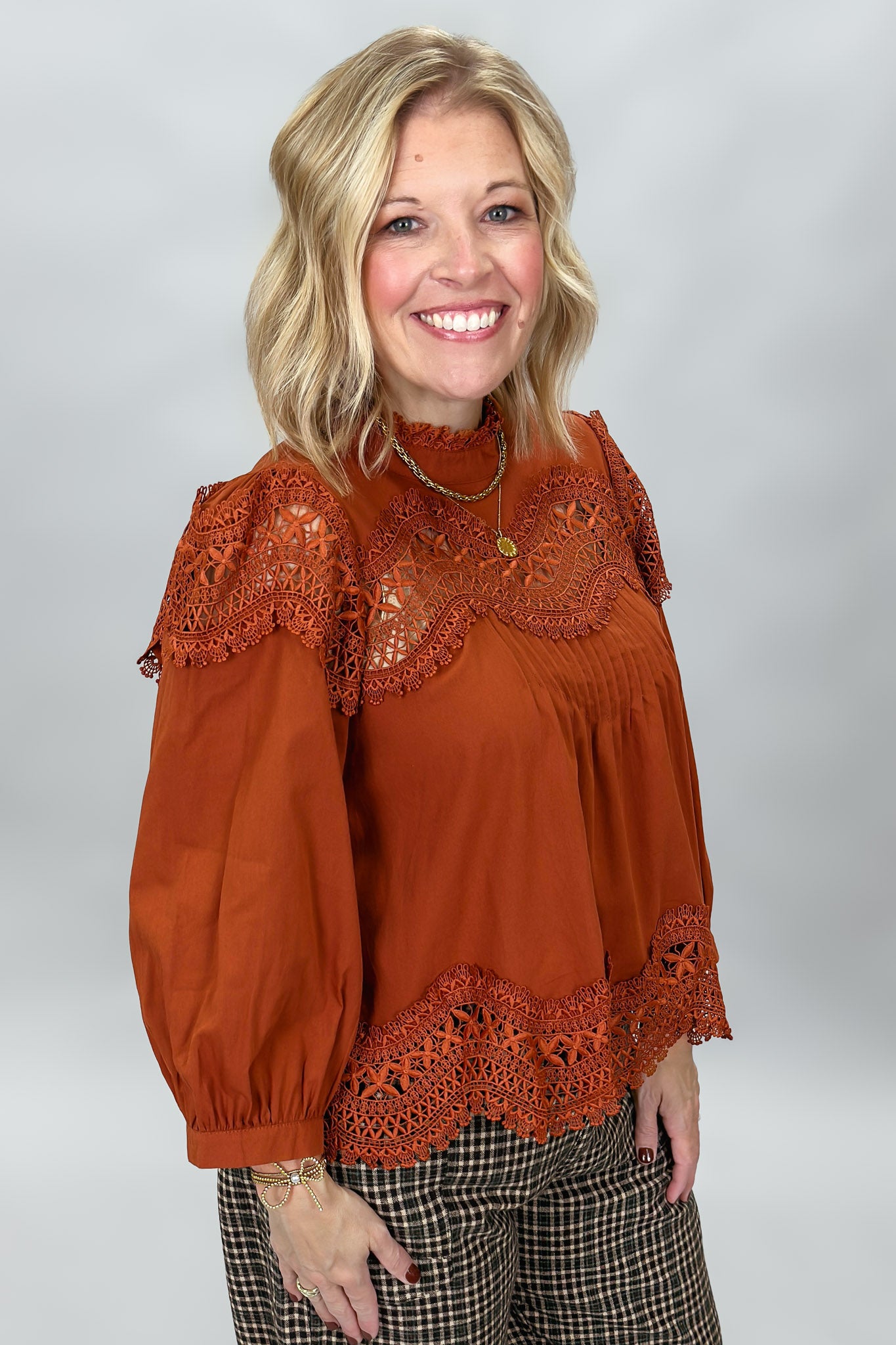 A smiling blonde woman wearing a rust-orange long-sleeve blouse with intricate lace trim at the neckline, cuffs, and hem, paired with brown and black plaid pants, against a neutral gray studio background.
