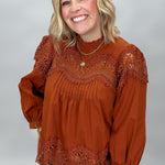 A smiling blonde woman wearing a rust-orange long-sleeve blouse with intricate lace and eyelet detailing across the neckline and sleeves, paired with black and white plaid pants, photographed against a neutral gray studio background.