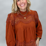 A smiling blonde woman wearing a rust-orange long-sleeve blouse with intricate lace detailing across the chest and hem, paired with brown and black plaid pants, photographed against a neutral gray studio background.
