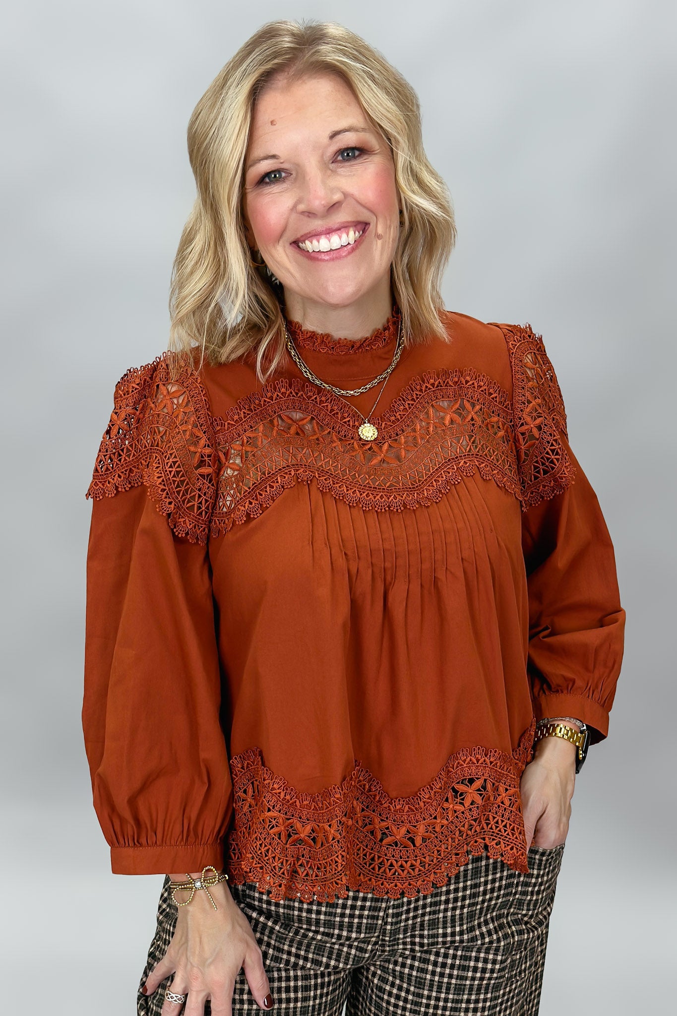 A smiling blonde woman wearing a rust-orange long-sleeve blouse with intricate lace detailing across the chest and hem, paired with brown and black plaid pants, photographed against a neutral gray studio background.