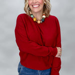 Phone, Keys, Wallet red relaxed bat-wing sleeve knit top, modeled with blue jeans and a colorful beaded necklace; front portrait showing the draped long sleeves and straight hem.