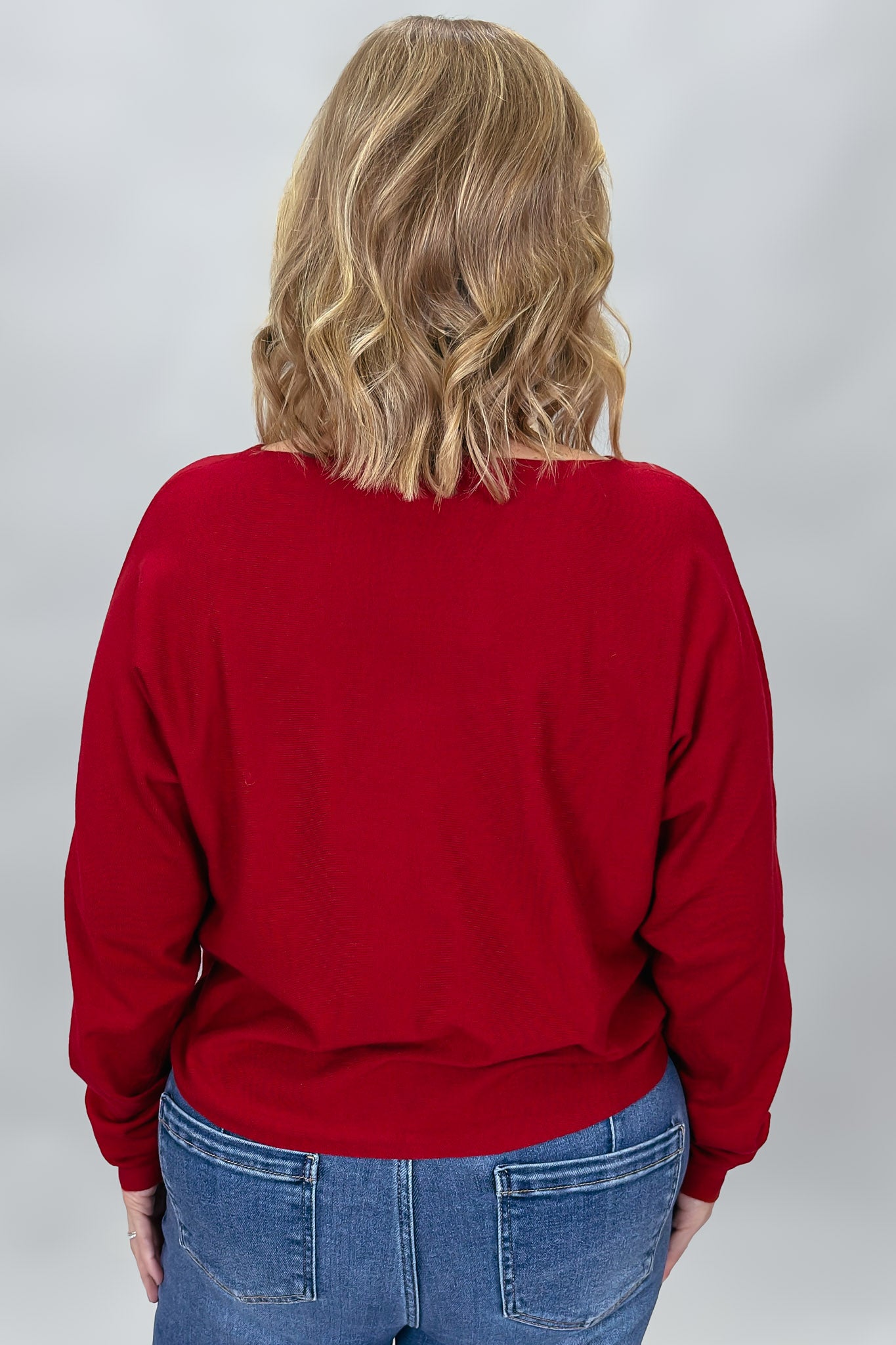 Back view of the Phone, Keys, Wallet red bat-wing sleeve sweater, showing its relaxed shoulder line, full sleeves, ribbed cuffs, and straight hem above blue jeans.