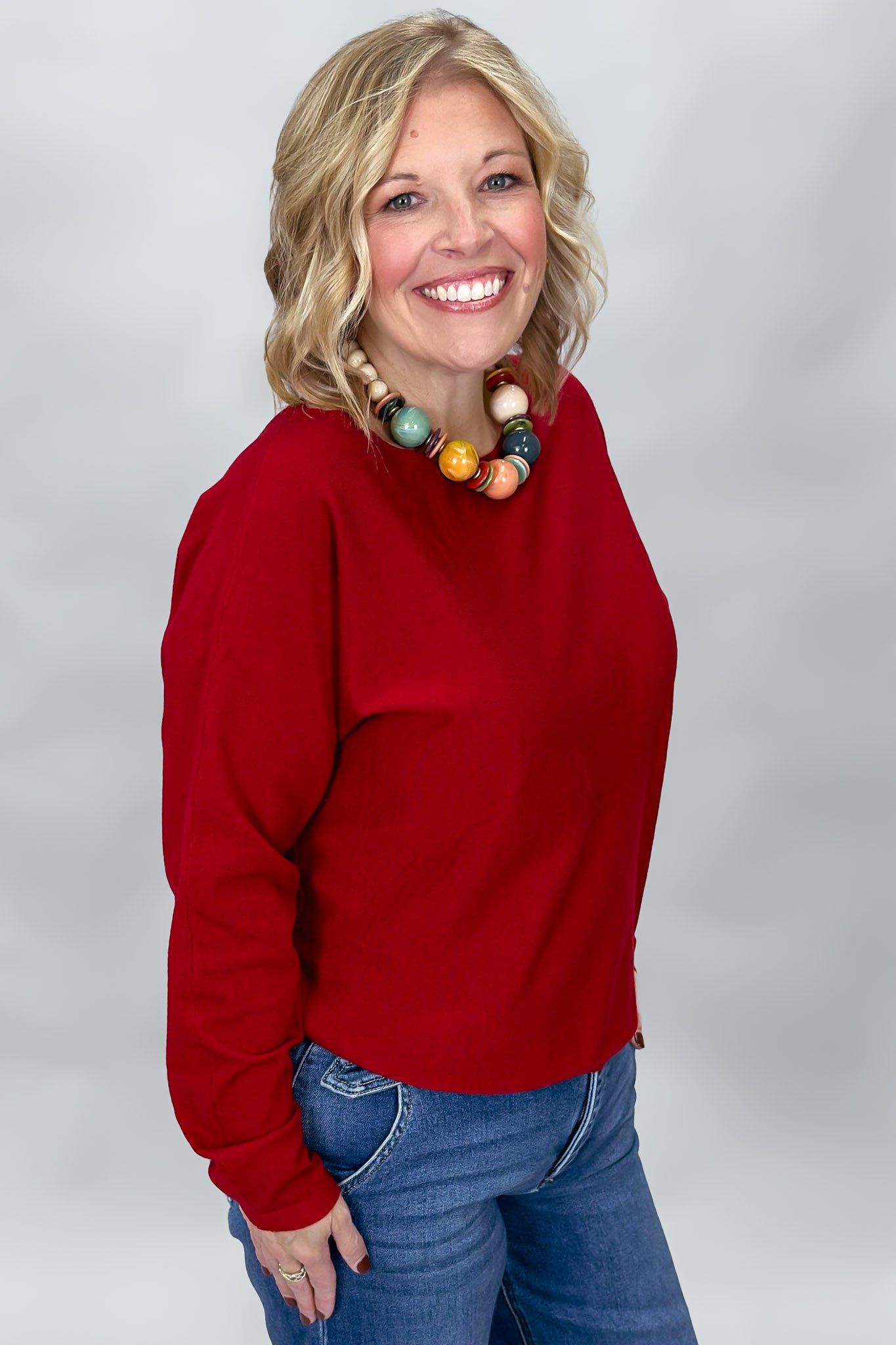 Phone, Keys, Wallet red relaxed knit top in a three-quarter model pose, showing the bat-wing sleeves, roomy body, hip-length hem, and casual fit over blue jeans.