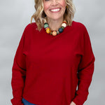 Phone, Keys, Wallet red bat-wing sleeve knit top in a straight-on waist-up view, featuring the crewneck, roomy fit, ribbed cuffs, and colorful beaded necklace over denim.