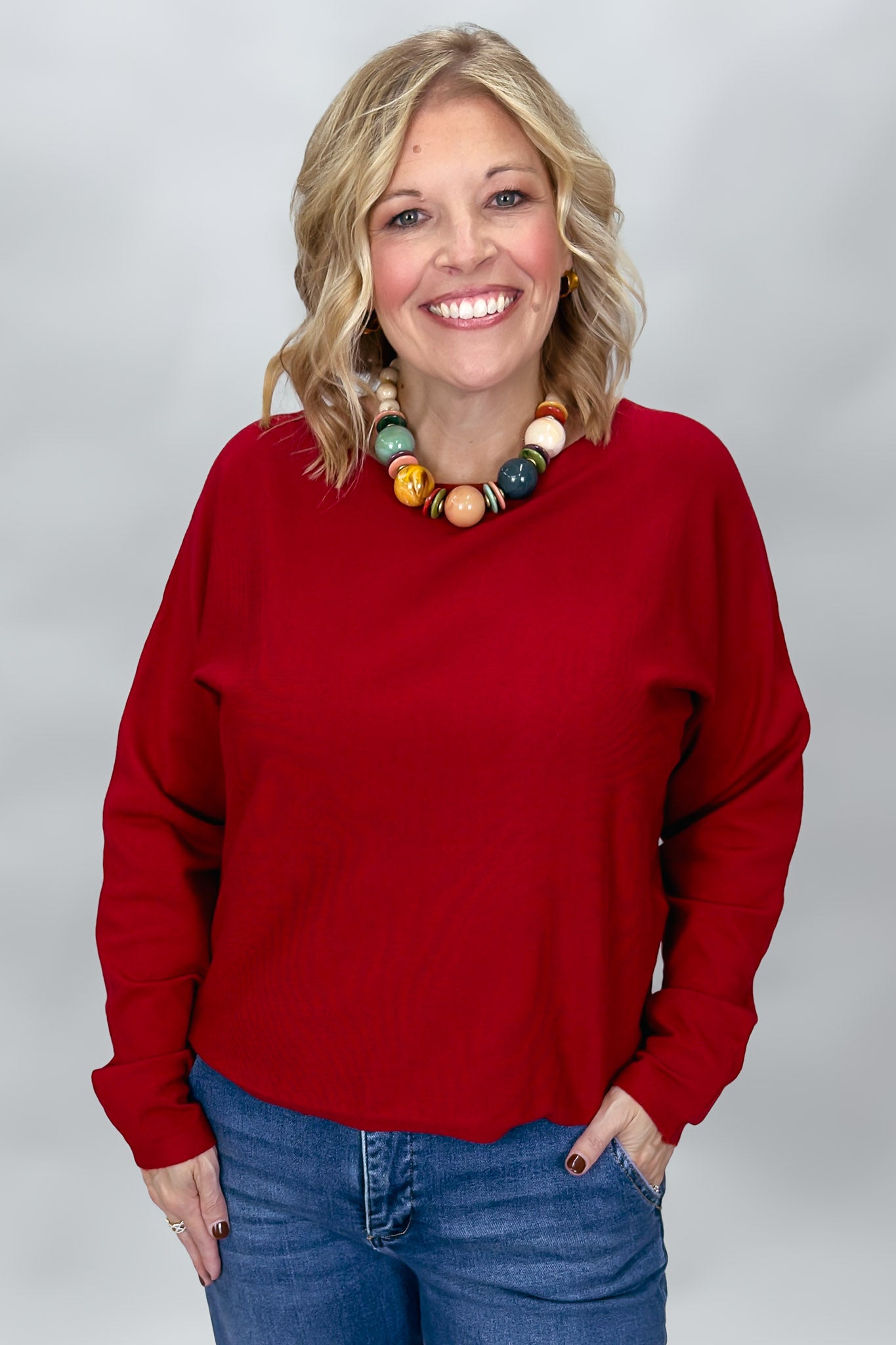 Phone, Keys, Wallet red bat-wing sleeve knit top in a straight-on waist-up view, featuring the crewneck, roomy fit, ribbed cuffs, and colorful beaded necklace over denim.