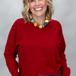 Phone, Keys, Wallet red bat-wing sleeve sweater close-up, showing the crew neckline, relaxed drape, ribbed cuffs, and soft knit texture with blue jeans and a wooden bead necklace.