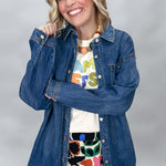 A smiling blonde woman poses in a studio wearing a blue denim button-up shirt over a white graphic t-shirt and a colorful floral skirt.