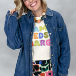 A smiling blonde woman models a blue denim button-up shirt over a cream graphic t-shirt and a colorful floral skirt against a neutral gray studio background.