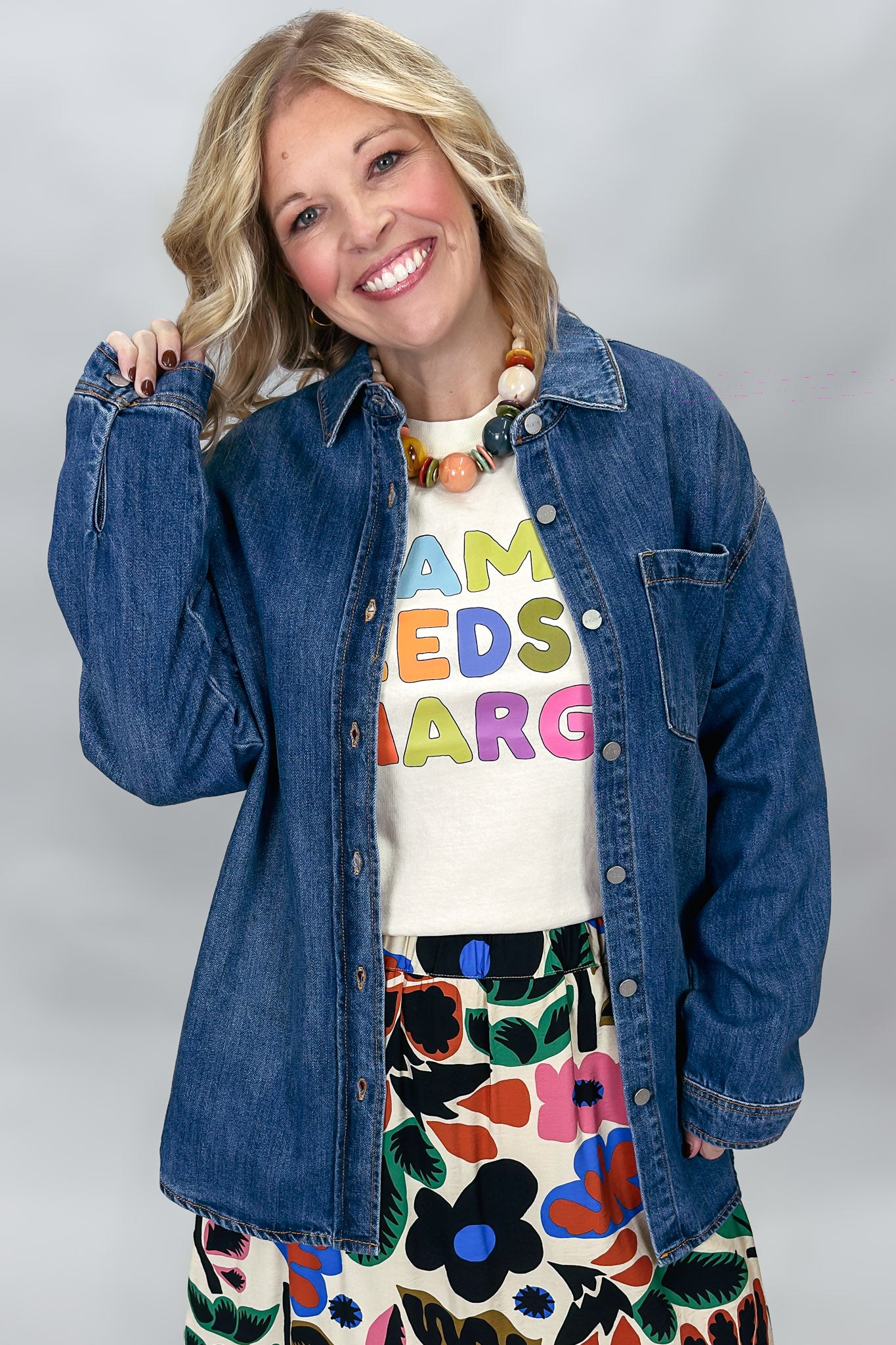 A smiling blonde woman models a blue denim button-up shirt over a cream graphic t-shirt and a colorful floral skirt against a neutral gray studio background.