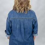 A woman viewed from behind wearing a blue denim shirt with rolled-up sleeves and a colorful patterned skirt, set against a light gray studio background.