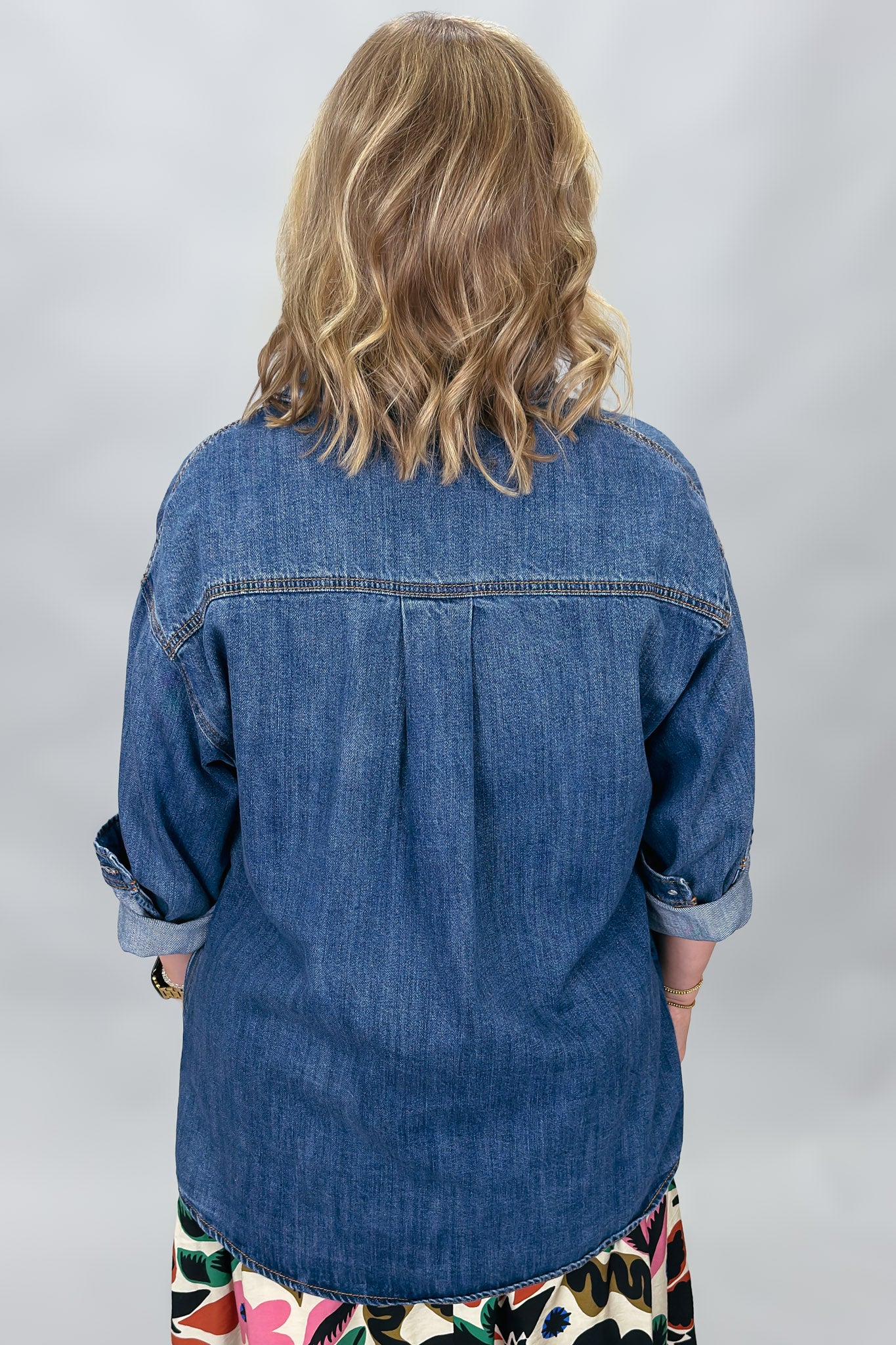 A woman viewed from behind wearing a blue denim shirt with rolled-up sleeves and a colorful patterned skirt, set against a light gray studio background.