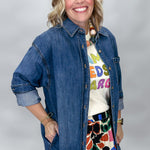 A smiling woman wearing a blue denim button-up shirt over a white graphic t-shirt and a colorful floral skirt, photographed against a light gray studio background.