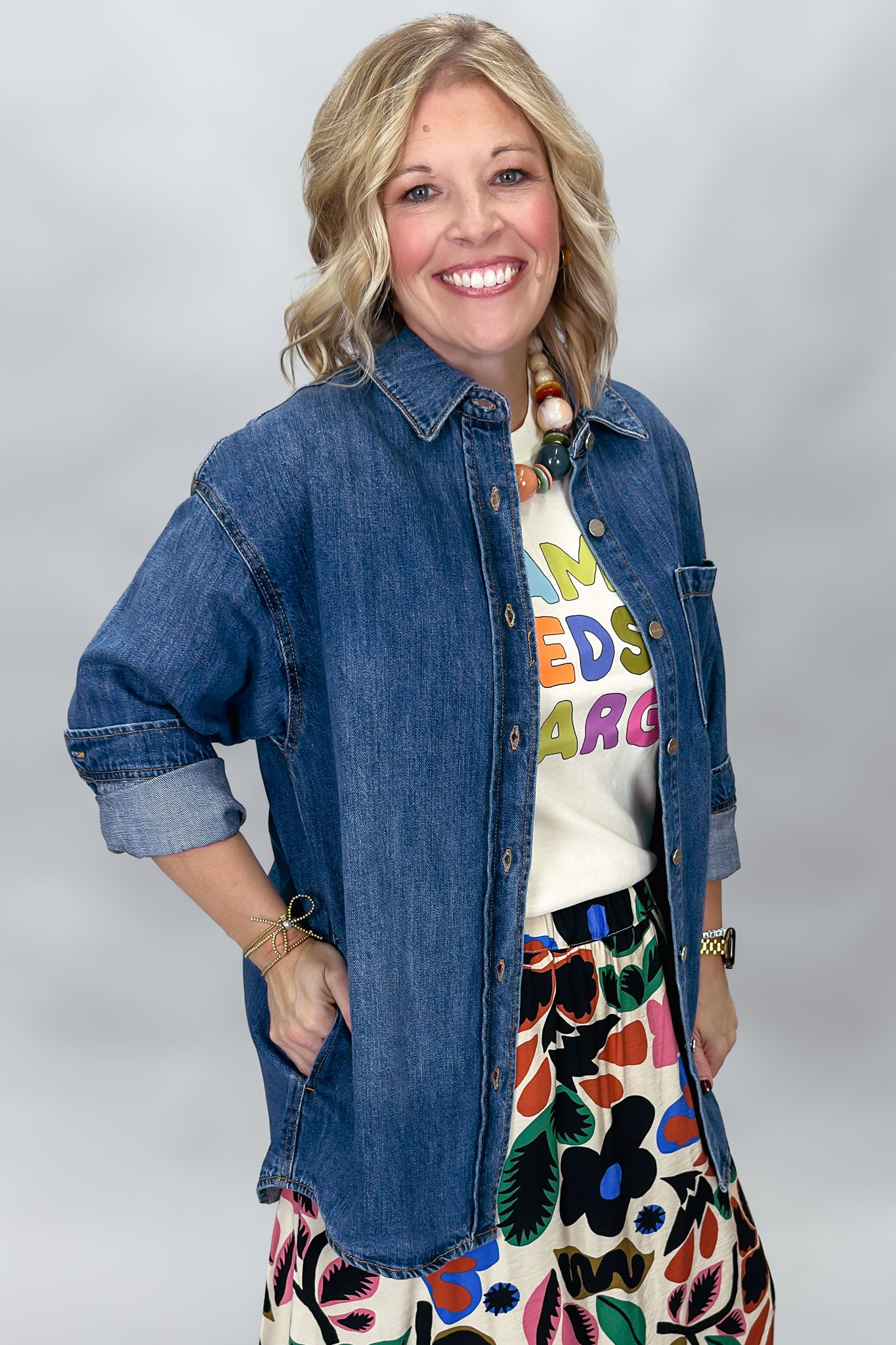 A smiling woman wearing a blue denim button-up shirt over a white graphic t-shirt and a colorful floral skirt, photographed against a light gray studio background.
