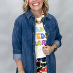 A smiling woman wearing a blue denim button-up shirt over a white graphic t-shirt and a colorful floral skirt, standing against a light gray studio background.