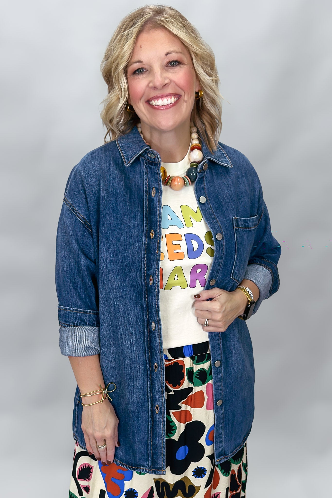 A smiling woman wearing a blue denim button-up shirt over a white graphic t-shirt and a colorful floral skirt, standing against a light gray studio background.