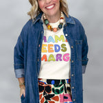 A smiling woman wearing a colorful graphic t-shirt under a blue denim shirt, accessorized with a chunky beaded necklace and a floral skirt, set against a plain gray studio background.