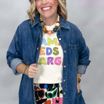 A smiling woman wearing a blue denim button-up shirt over a cream graphic T-shirt with colorful letters, paired with a colorful floral skirt, against a light gray studio background.