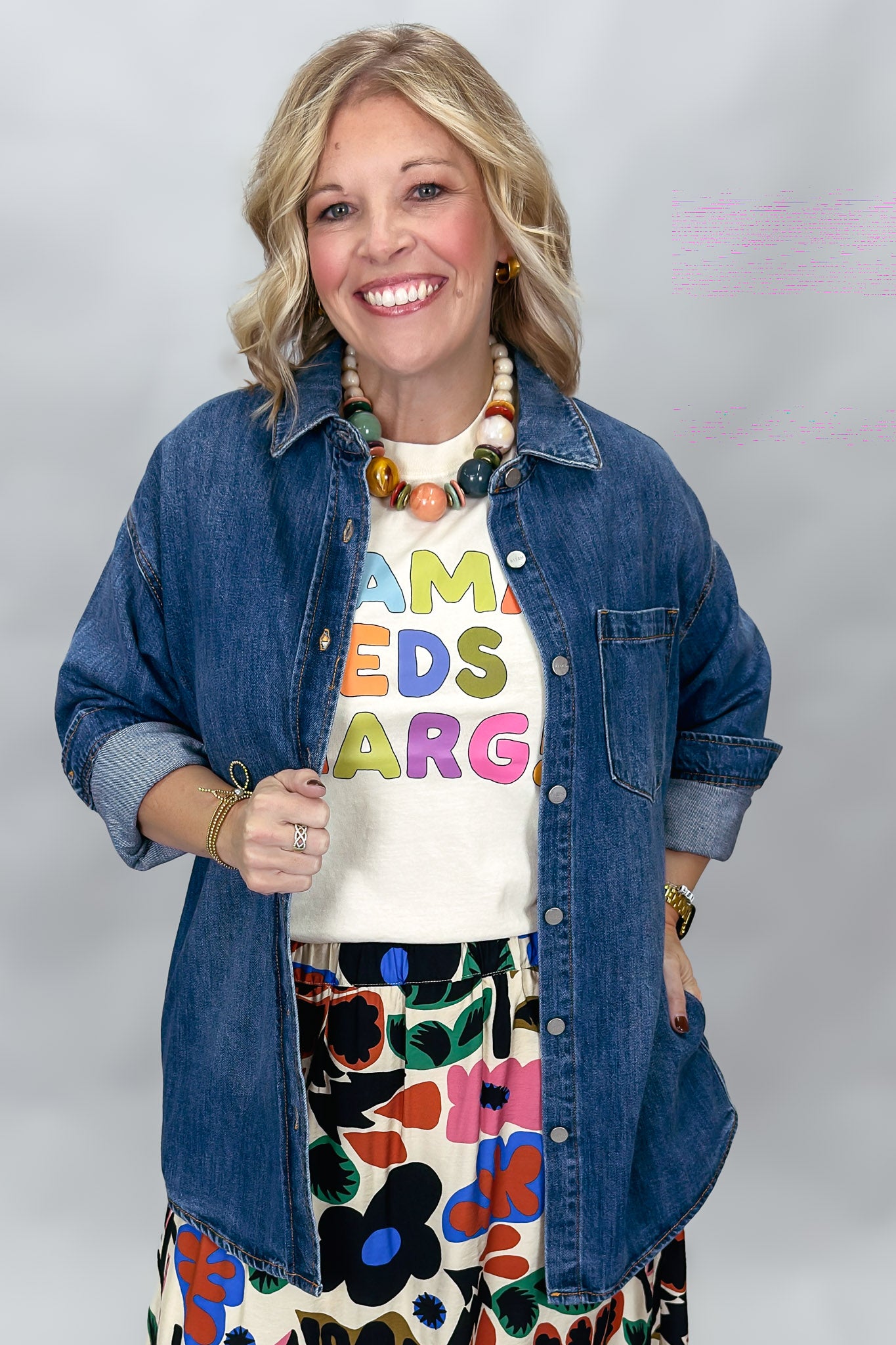 A smiling woman wearing a blue denim button-up shirt over a cream graphic T-shirt with colorful letters, paired with a colorful floral skirt, against a light gray studio background.