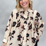 A smiling blonde woman wearing a cream-colored, smocked button-down blouse with a black and red floral pattern, paired with black pants, against a neutral gray studio background.
