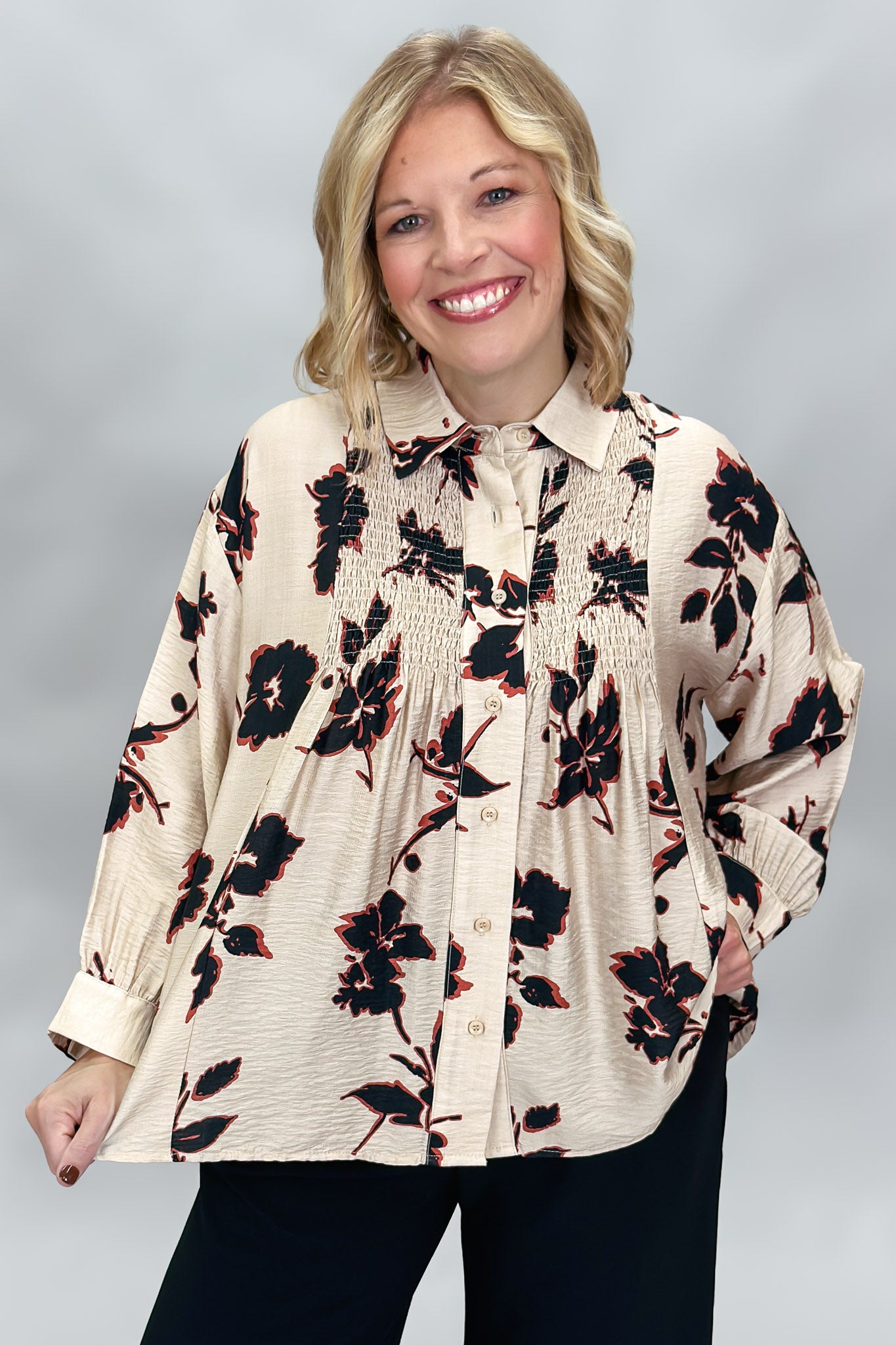 A smiling blonde woman wearing a cream-colored, smocked button-down blouse with a black and red floral pattern, paired with black pants, against a neutral gray studio background.