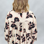 A woman viewed from behind wearing a cream-colored blouse with a black and red floral pattern, paired with dark pants, against a light gray studio background.