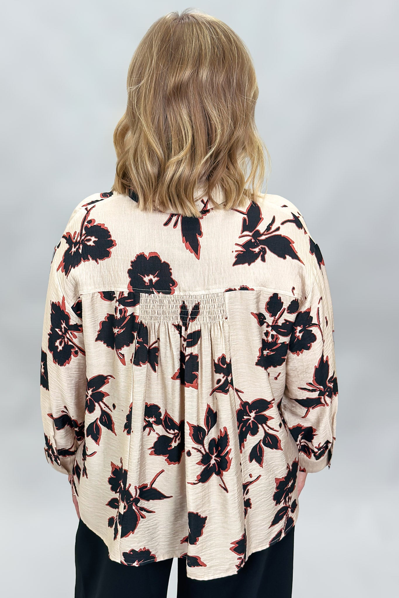 A woman viewed from behind wearing a cream-colored blouse with a black and red floral pattern, paired with dark pants, against a light gray studio background.