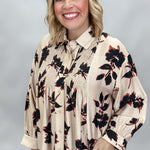 A smiling blonde woman wearing a beige long-sleeve blouse with a black and red floral print, paired with black pants, against a neutral gray studio background.