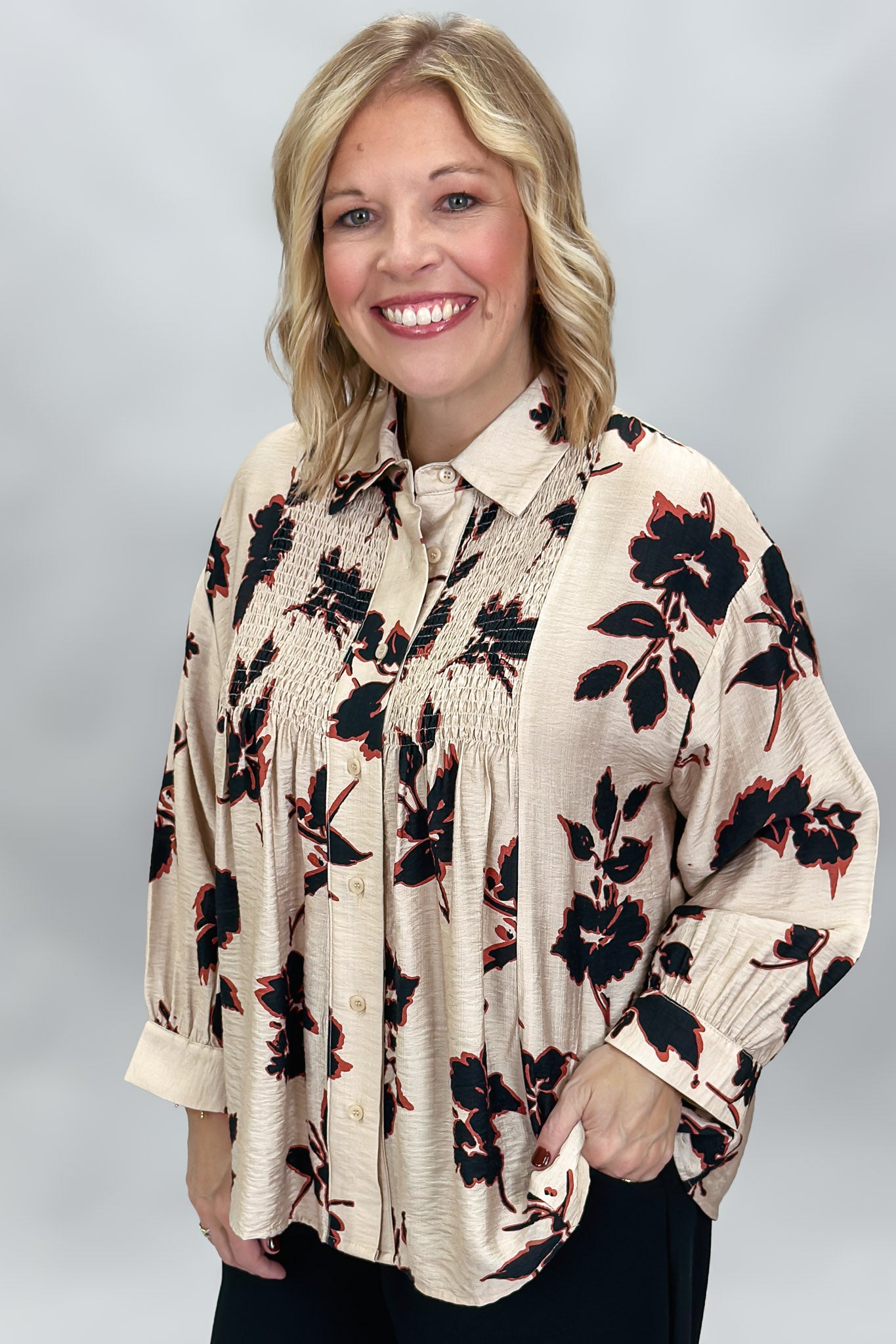A smiling blonde woman wearing a beige long-sleeve blouse with a black and red floral print, paired with black pants, against a neutral gray studio background.