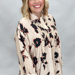 A smiling blonde woman wearing a beige long-sleeve blouse with a black and red floral pattern, paired with black pants, against a neutral gray studio background.