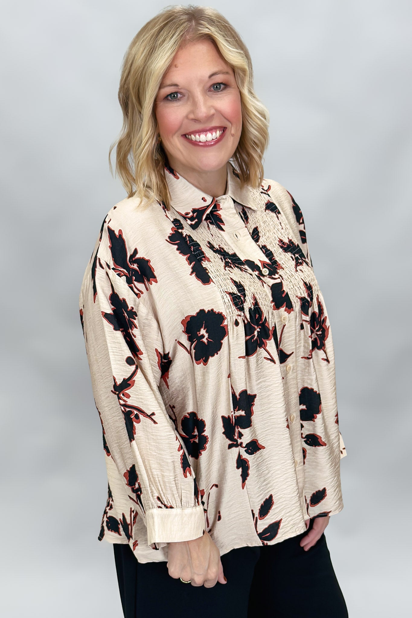 A smiling blonde woman wearing a beige long-sleeve blouse with a black and red floral pattern, paired with black pants, against a neutral gray studio background.