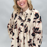A smiling blonde woman wearing a beige long-sleeve blouse with a black and red floral pattern, paired with black pants, photographed against a neutral gray studio background.