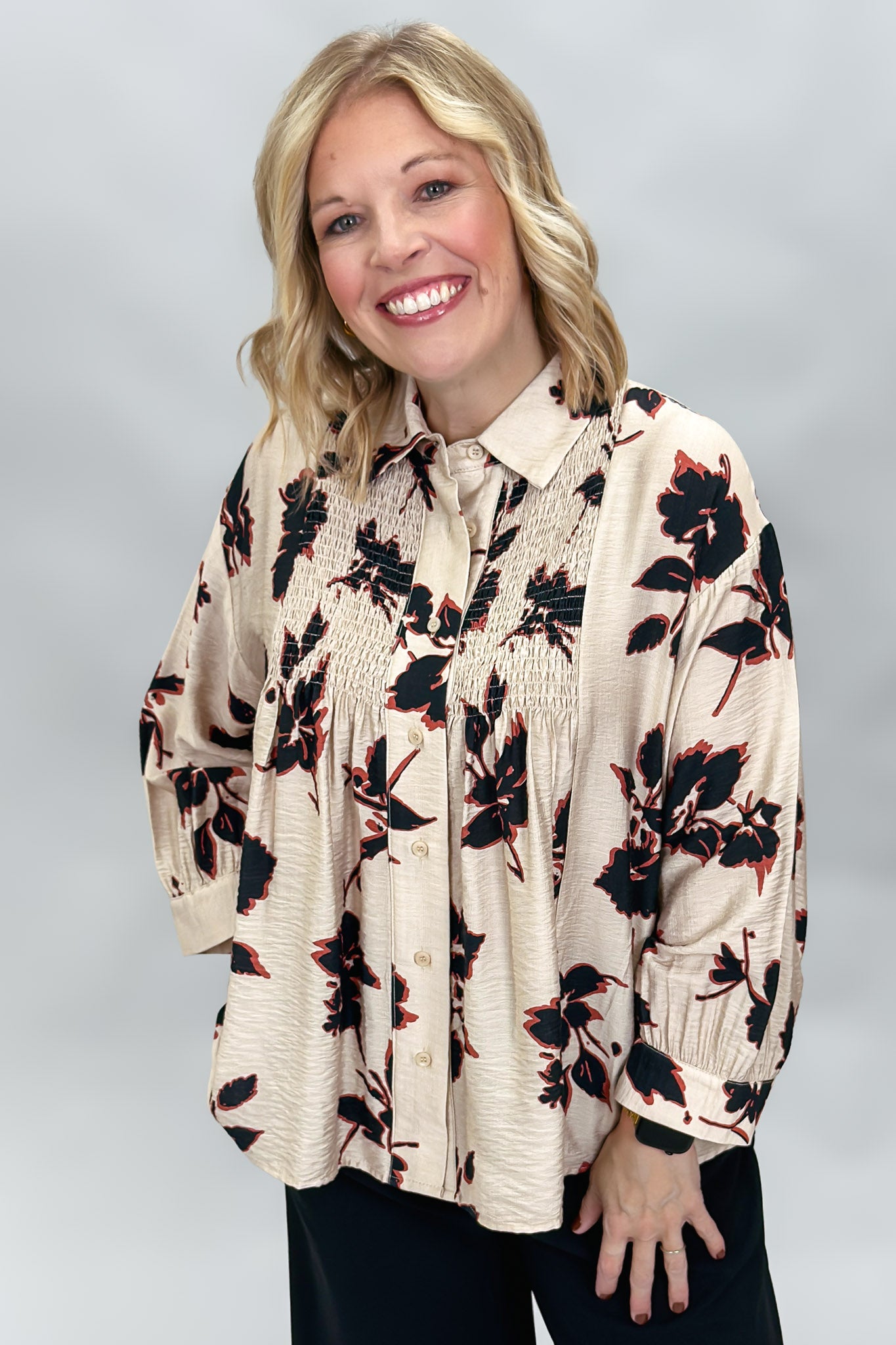 A smiling blonde woman wearing a beige long-sleeve blouse with a black and red floral pattern, paired with black pants, photographed against a neutral gray studio background.